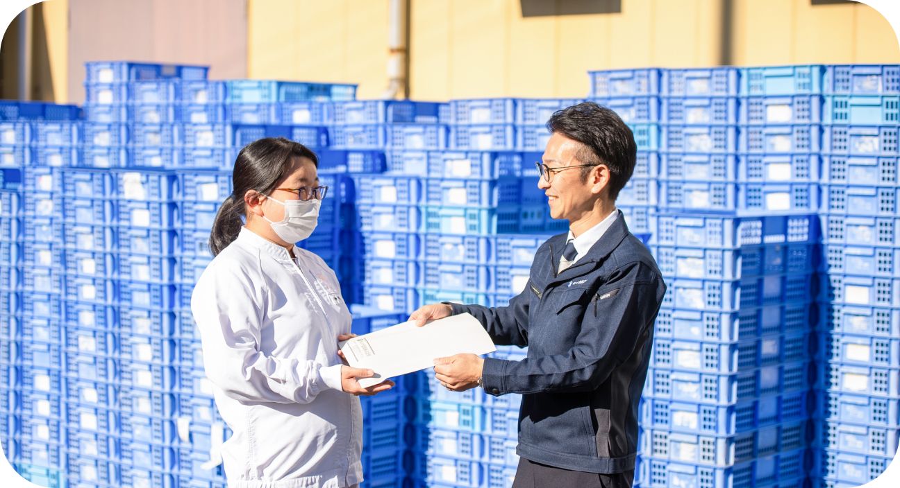 青い物流コンテナが積まれた屋外で書類を手渡し合う女性社員と男性社員