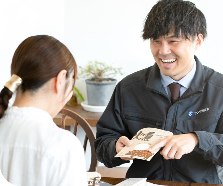 サンワ包装のロゴ入り作業着姿で、女性のお客様に笑顔でパッケージ商品を提案する営業社員
